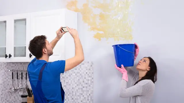 Man photographing a ceiling water stain while a woman holds a bucket